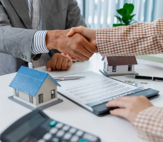 Two people in business attire finalize a real estate deal, shaking hands across a desk with house models, keys, and a contract
